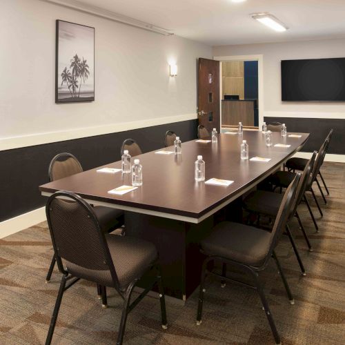 A conference room with a long table, chairs, water bottles, notepads, a framed picture, and a TV screen on the wall.