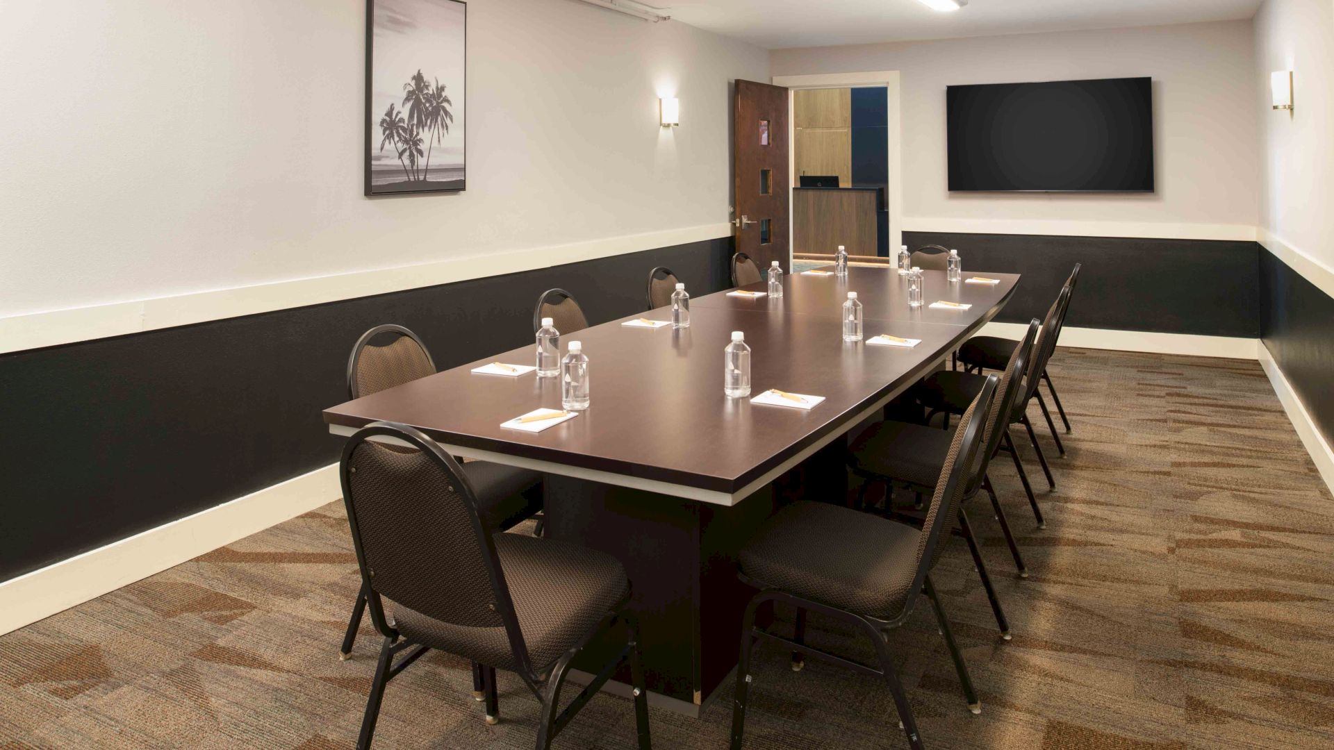 A conference room with a long table, chairs, water bottles, and a TV. There&rsquo;s a framed picture on the wall and a carpeted floor.