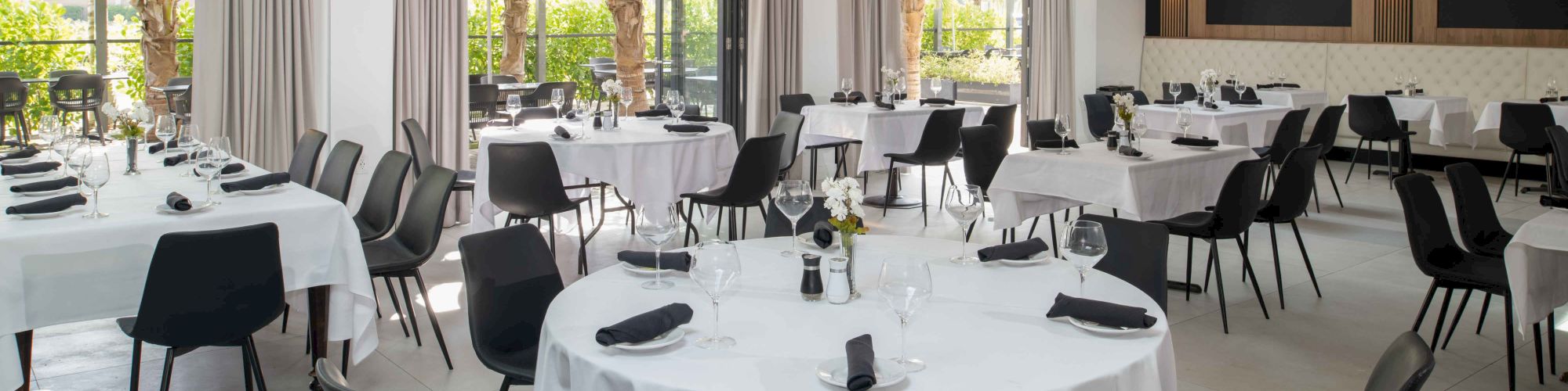 A spacious, elegantly set dining room with round tables, white tablecloths, and black chairs, ready for guests. Large windows offer a scenic view.