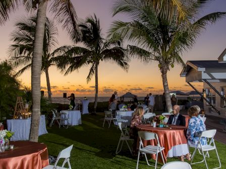 Outdoor event with tables, chairs, and palm trees at sunset. The scene suggests a relaxed, tropical gathering.