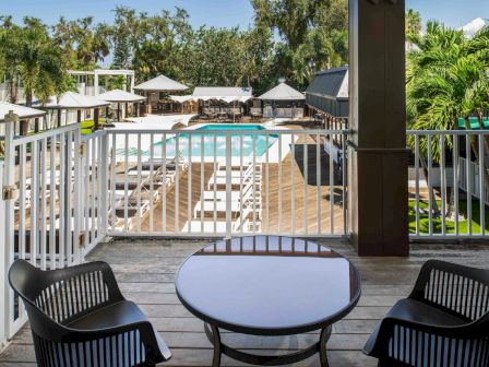 A balcony with a table and two chairs overlooks a pool area surrounded by loungers and palm trees, creating a relaxing outdoor atmosphere.