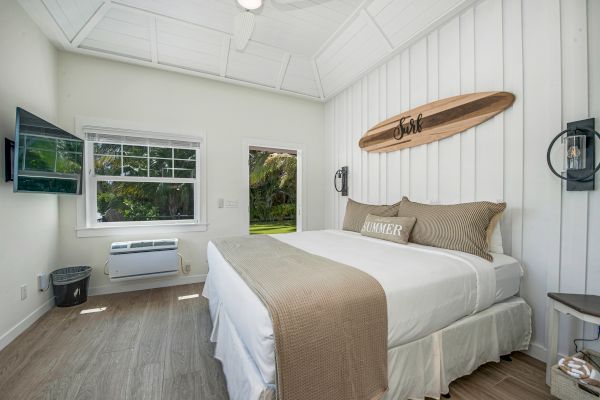 A cozy bedroom with a large bed, surfboard decor, TV, window, and door leading outside. Light wood flooring and neutral tones complete the look.