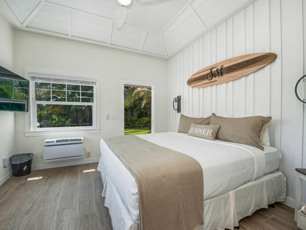 A cozy bedroom with a large bed, surfboard decor, TV, window, and door leading outside. Light wood flooring and neutral tones complete the look.