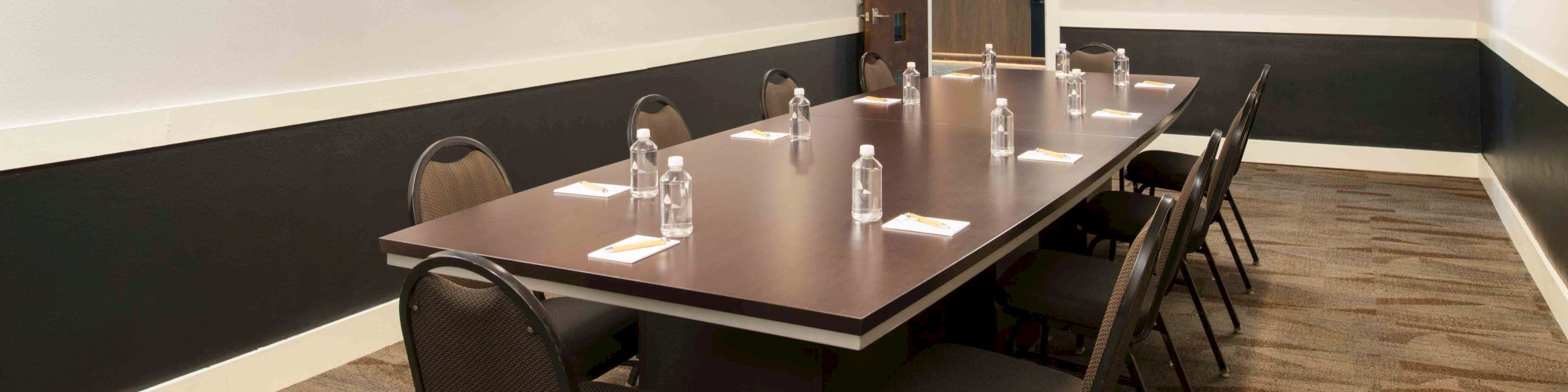 A conference room with a long table, chairs, water bottles, and a TV. There’s a framed picture on the wall and a carpeted floor.