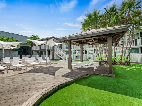 The image shows an outdoor lounge area with sun loungers, umbrellas, a wooden deck, and a pergola, surrounded by palm trees and greenery.