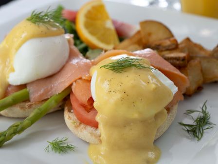 Eggs Benedict with poached eggs, hollandaise sauce, smoked salmon, asparagus, tomato, and a side of potatoes and orange slice.