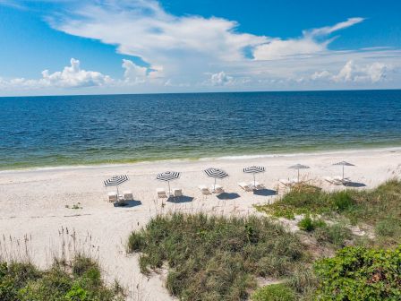 A serene beach scene with white sand, several umbrellas, lush greenery, calm ocean, and a partly cloudy blue sky in the background.