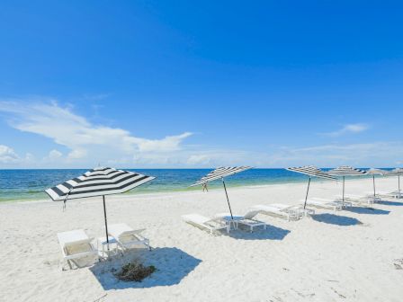 A beach scene with striped umbrellas and lounge chairs on white sand, facing a blue ocean under a clear sky with a few clouds.