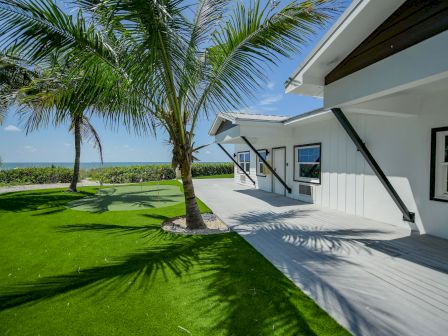 A modern house with a gray deck, surrounded by green grass and palm trees, overlooking the ocean under a clear blue sky.