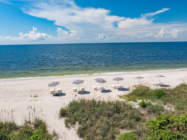 A serene beach scene with white sand, several umbrellas, lush greenery, calm ocean, and a partly cloudy blue sky in the background.
