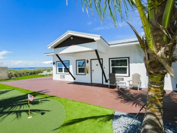 A modern house with a wooden deck, adjacent patio furniture, palm tree, and a mini-golf area overlooking the ocean.