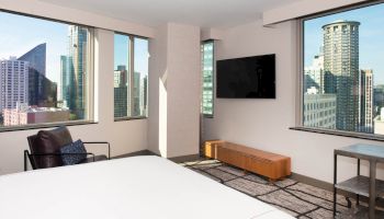 The image shows a modern hotel room with large windows offering a cityscape view, a bed, an armchair, and a wall-mounted TV.