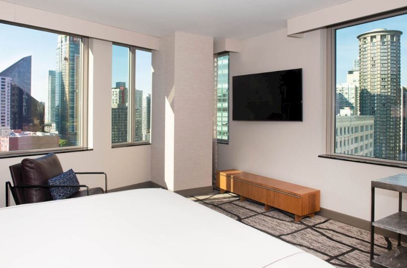 The image shows a modern hotel room with large windows offering a cityscape view, a bed, an armchair, and a wall-mounted TV.