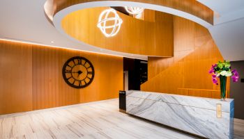 The image shows a modern indoor area with a marble reception desk, a large wall clock, wooden paneling, and decorative ceiling lights.