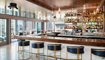 The image shows a modern bar with shelves of liquor bottles, bar stools, a wooden countertop, and large windows providing a cityscape view.