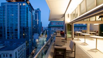 A cityscape at dusk with high-rise buildings and a modern, well-lit balcony featuring tables, chairs, and large windows on the right side.