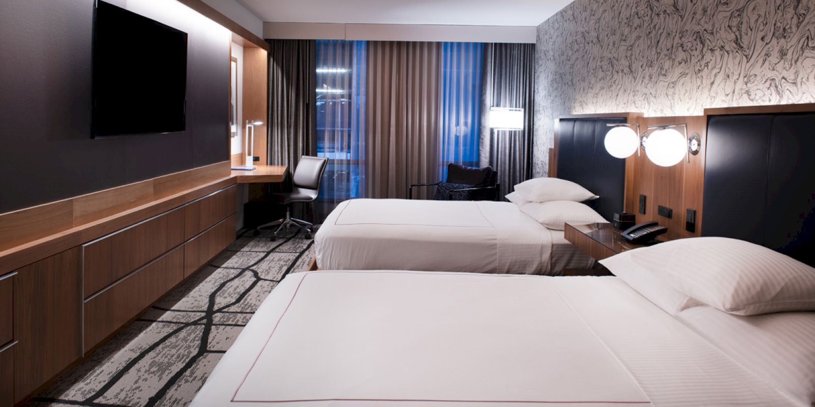 This image shows a hotel room with two beds, a wall-mounted TV, a desk with a chair, and modern decor featuring a patterned wall and long curtains.