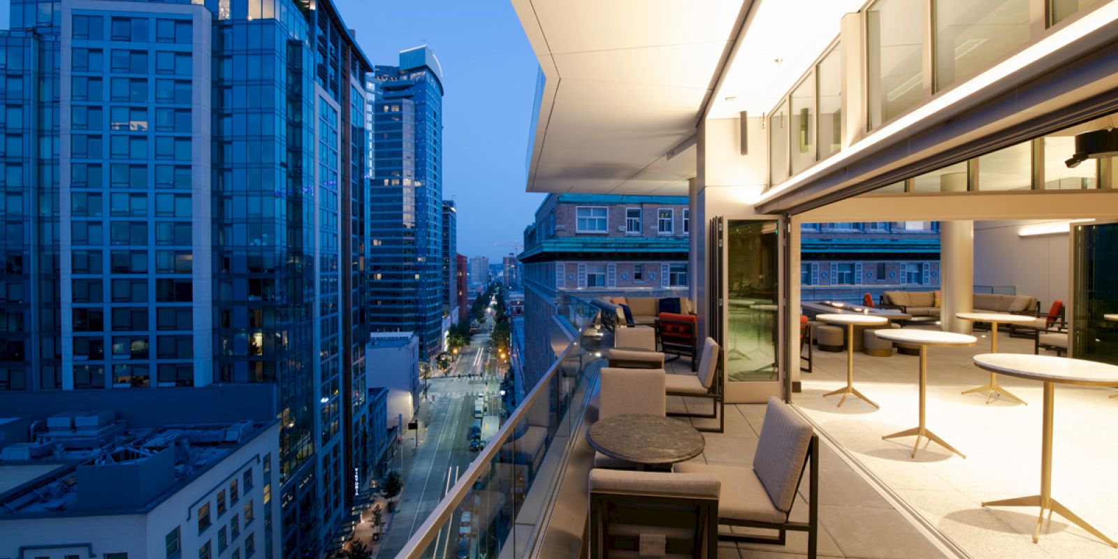This image shows a cityscape with tall buildings at dusk, alongside a modern balcony with tables and chairs.