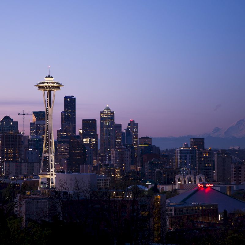 A cityscape at dusk, featuring a prominent tower and downtown skyline with a mountain in the background, under a vibrant evening sky.