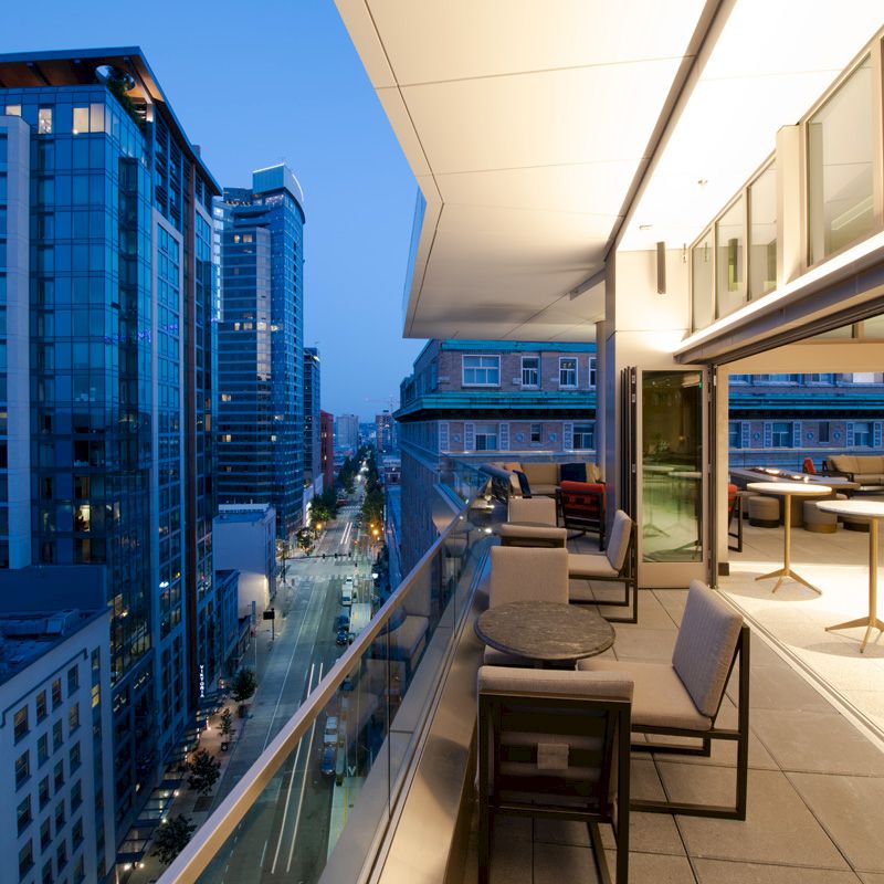 An elevated outdoor terrace with seating overlooks a city street lined with modern buildings, illuminated as evening approaches.