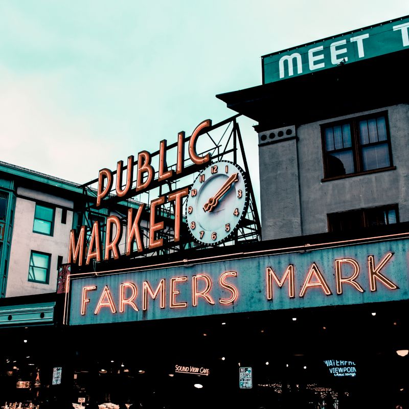An image of the iconic Public Market sign at Pike Place Market in Seattle. A clock and "Farmers Market" neon sign are also visible.