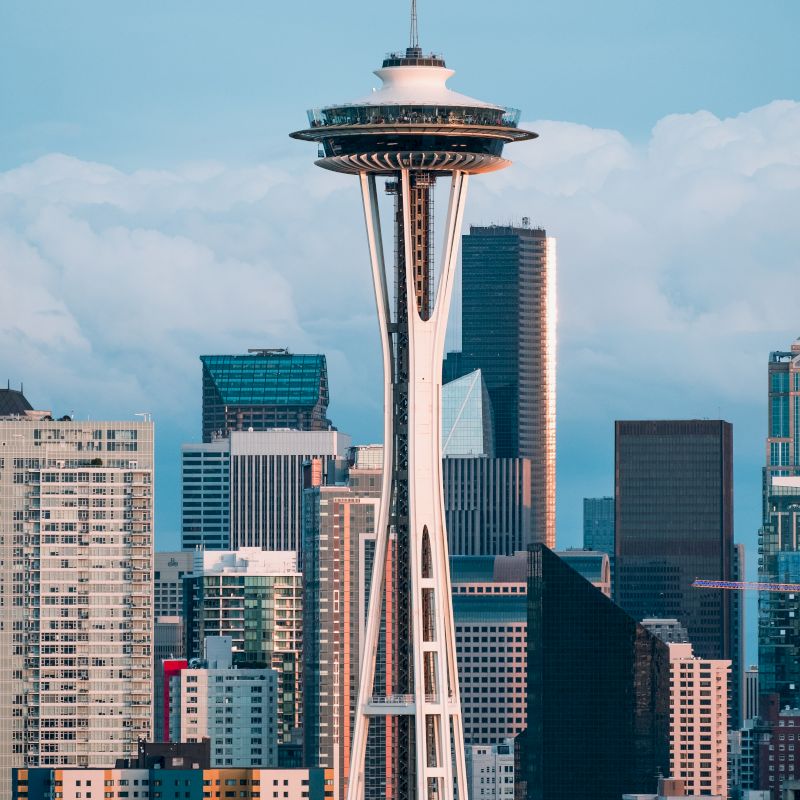 The image shows the Space Needle surrounded by various high-rise buildings in Seattle, Washington, with a partly cloudy sky in the background.