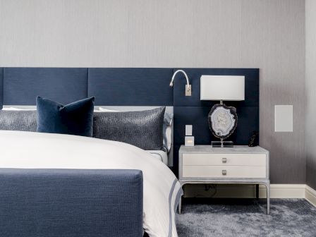 A modern bedroom with a navy and white theme, featuring a bed with blue pillows, a nightstand with a lamp, and a decorative object on it.
