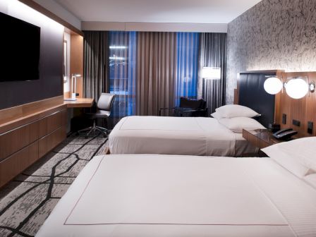 The image shows a modern hotel room with two twin beds, a television mounted on the wall, a work desk, and large windows with curtains.