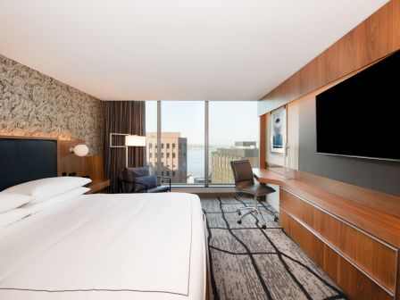 A modern hotel room features a bed, a desk with a chair, a large TV, and a window with a city view.