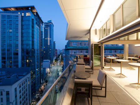 A modern cityscape at dusk, featuring a sleek balcony with seating and tables overlooking a street lined with tall buildings and lights.