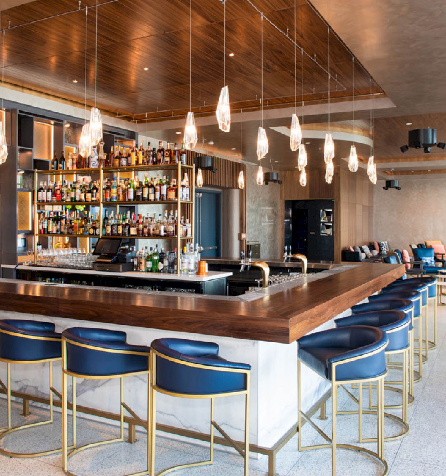 A stylish bar with modern decor features a long counter with blue chairs, hanging lights, and a well-stocked shelf of various bottles.