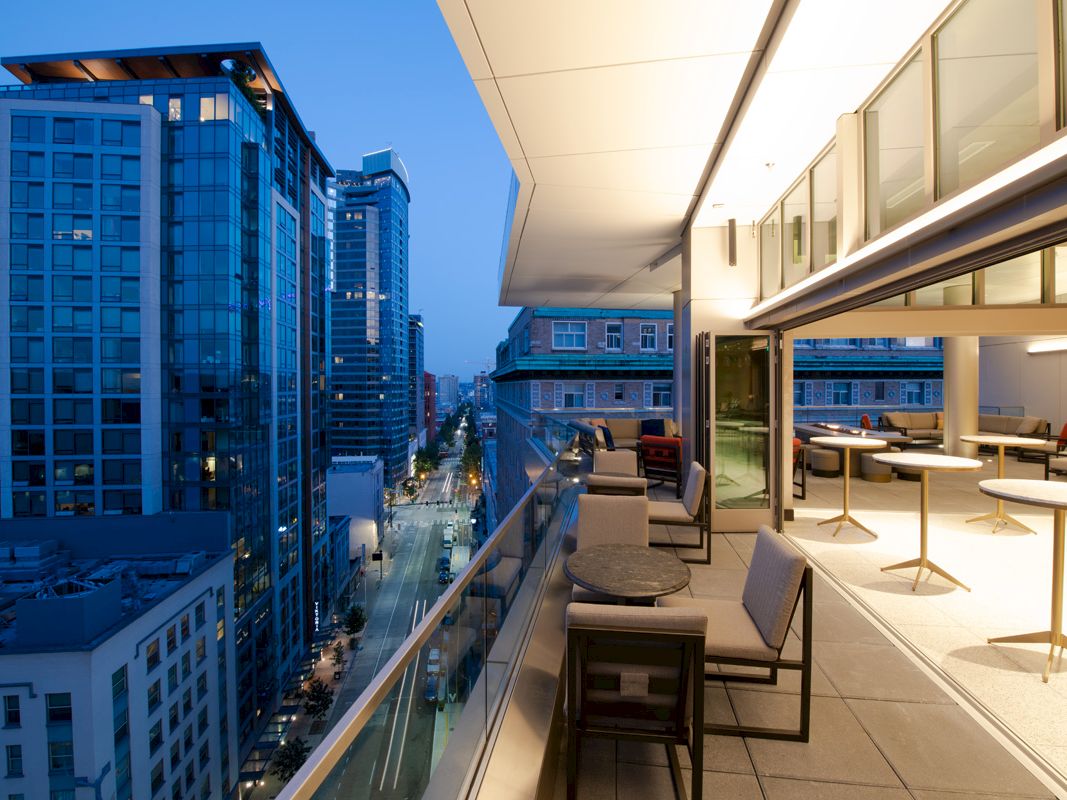 A modern balcony overlooking a cityscape with tall buildings, featuring seating areas, small tables, and glass railings during the evening.