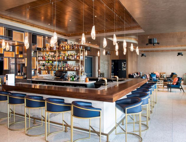 The image shows a well-designed bar with a central counter and high stools, surrounded by a cozy seating area and modern decor elements.