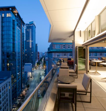 A modern cityscape at dusk features a stylish balcony with tables and chairs, overlooking high-rise buildings along a well-lit street.