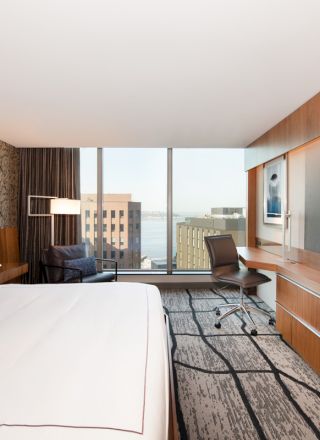 A modern hotel room features a large bed, wall art, a flat-screen TV, a work desk, a chair by the window, and a view of buildings outside.