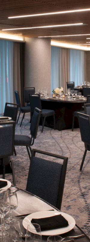 The image shows a dimly lit banquet hall with round tables set for an event, featuring black chairs, white floral centerpieces, and neatly arranged tableware.