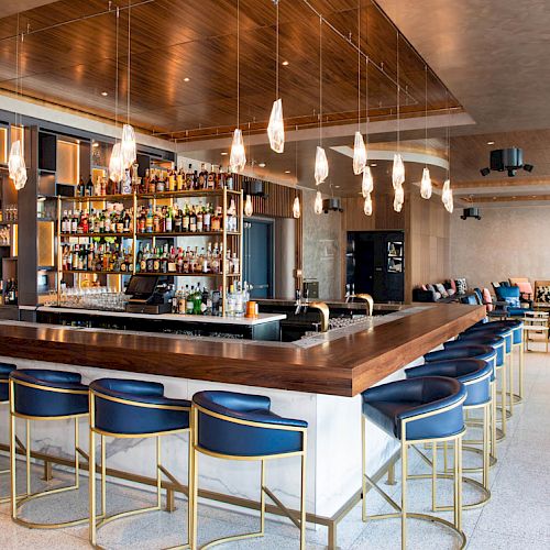 A modern bar with elegant lighting, blue bar stools, a well-stocked bar counter, and a stylish seating area with a mix of chairs and sofas.