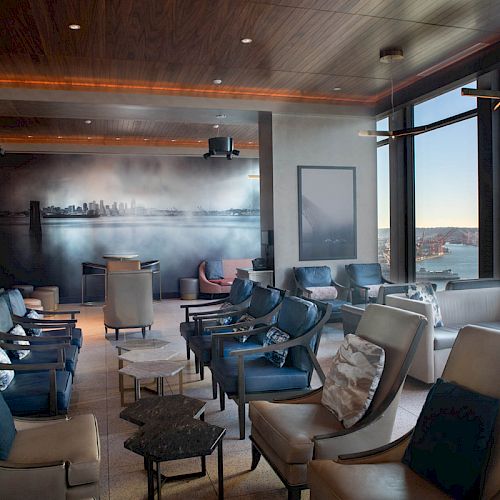 A modern lounge with numerous seats, large windows offering scenic water views, and contemporary decor featuring a city skyline mural on the wall.