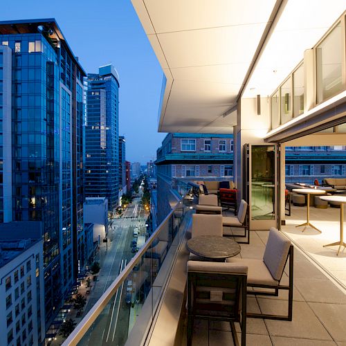 A modern cityscape at dusk, featuring high-rise buildings and a rooftop terrace with seating and tables overlooking a bustling street.