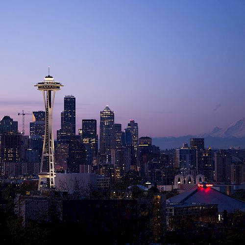 The image shows a city skyline at dusk featuring a distinctive tower, with mountains in the background and various buildings illuminated.