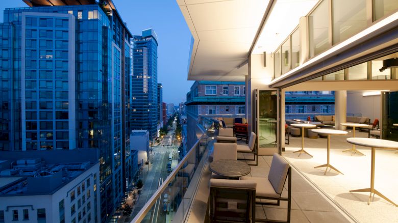 An urban balcony view with seating and tables, overlooking a city street and modern high-rise buildings, taken during evening time.