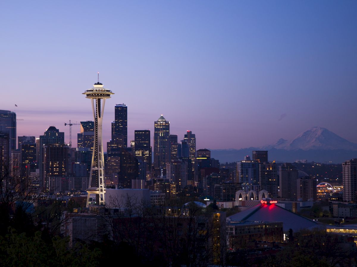 A cityscape featuring the Space Needle, surrounded by modern buildings at dusk with a distant mountain in the background, captured under a twilight sky.