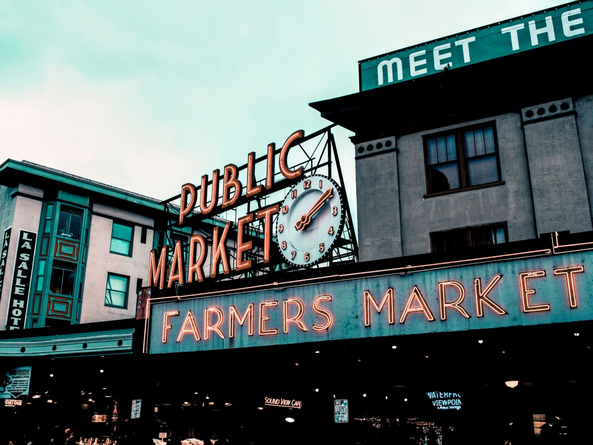 The image shows an iconic "Public Market" sign above a "Farmers Market" sign with a clock in between, likely depicting Pike Place Market in Seattle.