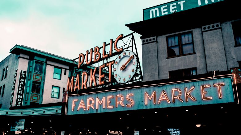 The image shows an iconic "Public Market" sign above a "Farmers Market" sign with a clock in between, likely depicting Pike Place Market in Seattle.