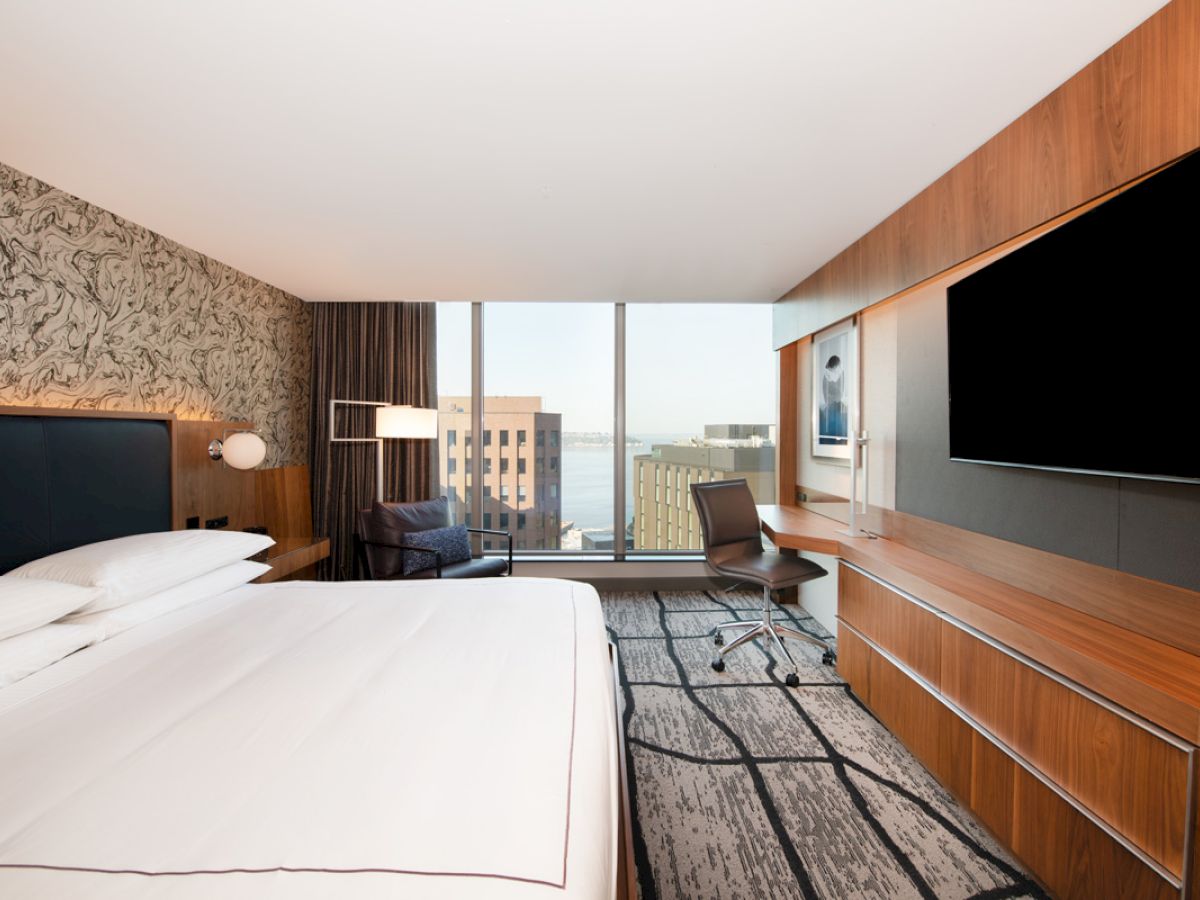 A modern hotel room with a large bed, wall-mounted TV, desk, chair, and window with a city view in the background.
