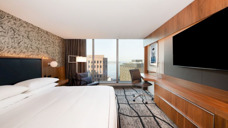 A modern hotel room with a large bed, wall-mounted TV, desk, chair, and window with a city view in the background.