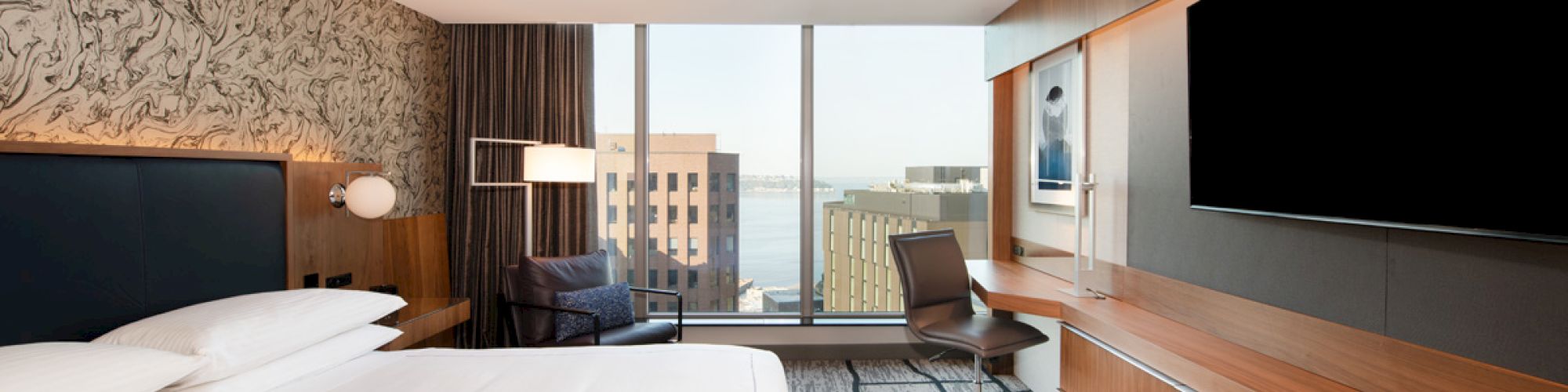 A modern hotel room features a large bed, a flat-screen TV, a desk, and a window with a city view, including various buildings.